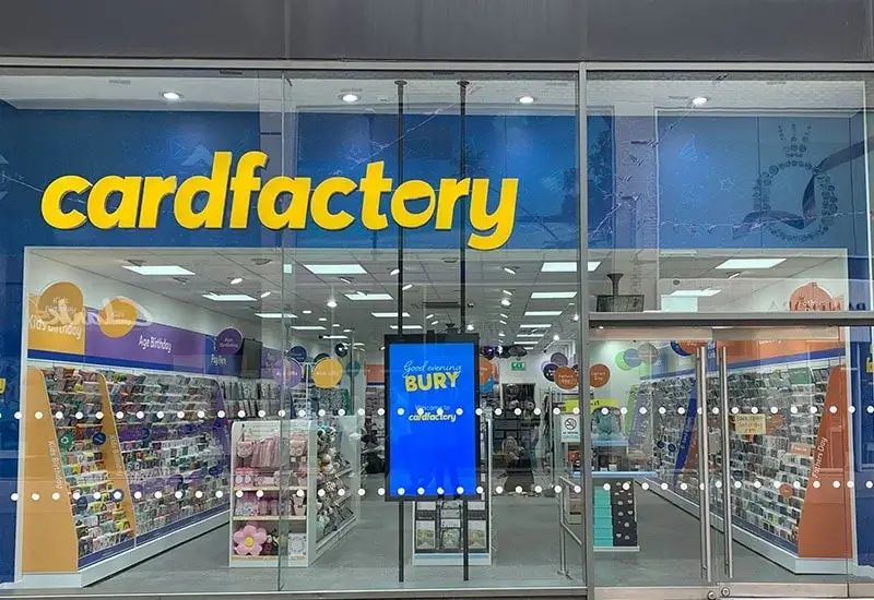Storefront of a 'cardfactory' shop with a glass facade, bright yellow 'cardfactory' logo on a blue background, shelves of greeting cards and gifts inside, and a digital sign reading 'Good morning BURY'
